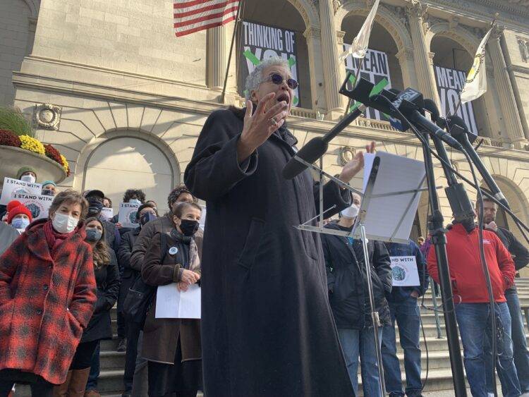 Toni Preckwinkle, dressed in a long black coat with circulars sunglasses, speaks to a line of microphones. Her mouth is open and her hands are raised as if in disbelief. 
