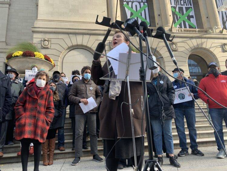 A woman in a long brown coat stands at a microphone stand addressing a crowd out of view. Behind her is a group of union supporters and the Art Institute of Chicago.