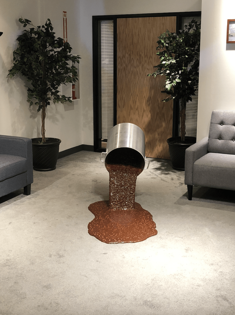 A sculpture of chili spilling from a metal pot inside of a bland office.