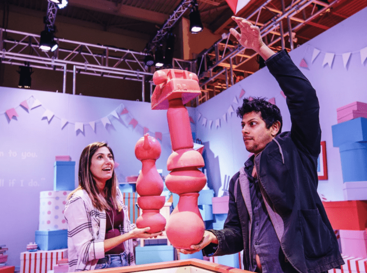 A man and woman balance pink-red objects at an interactive station; around them are pink, blue, and red boxes and banners.
