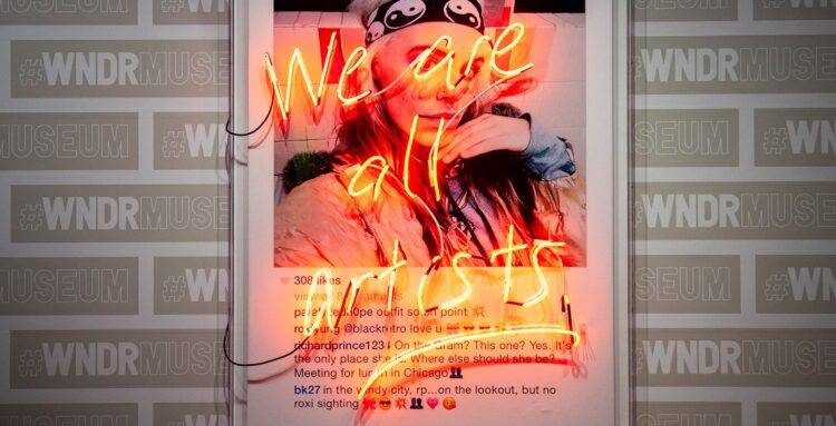 A large print-out of an Instagram post overlaid with neon lights shaped to read "We Are All Artists."
