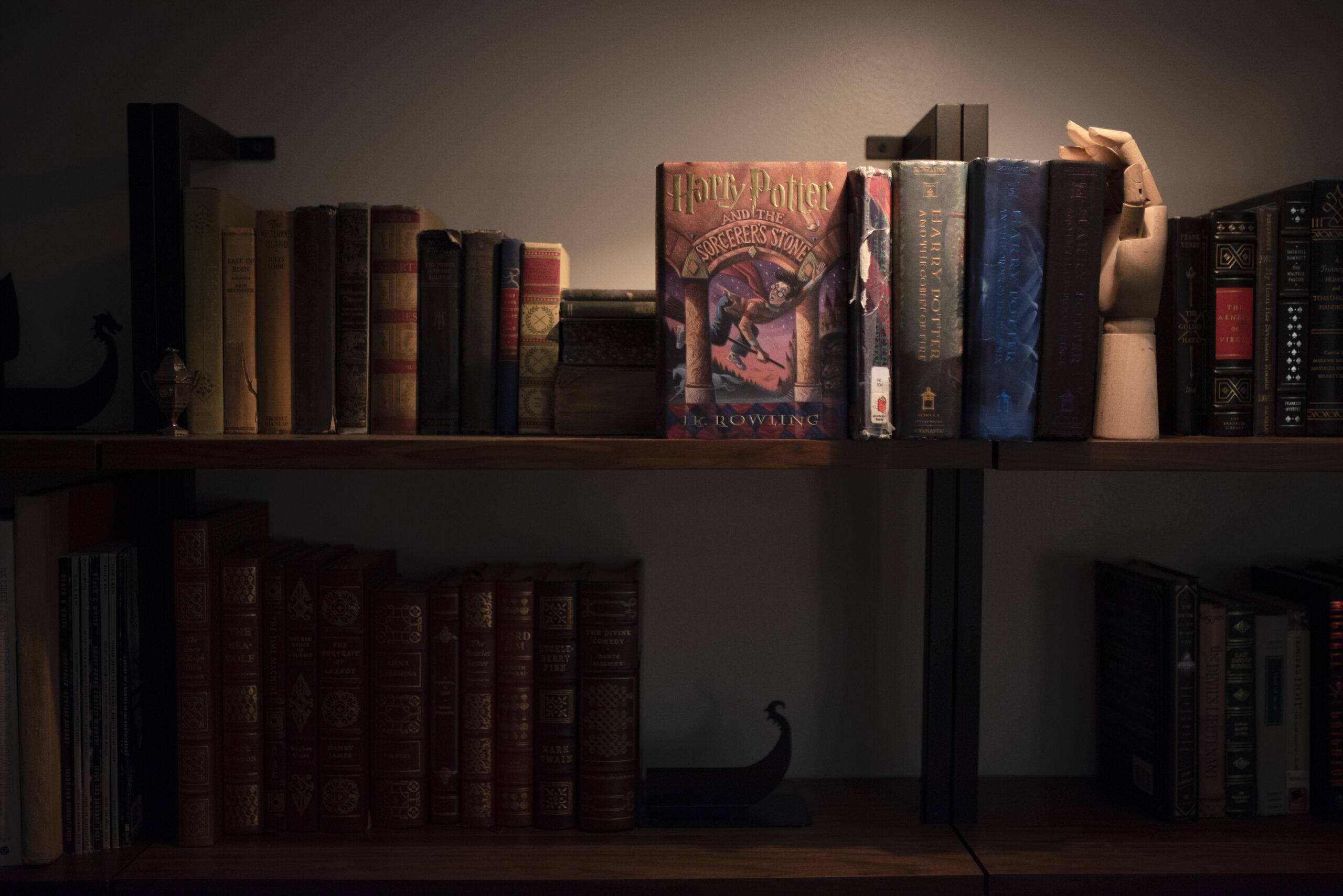 Rows of books on a bookshelf cast in shadow, a single spotlight illuminates several books from J.K. Rowling's "Harry Potter" series.