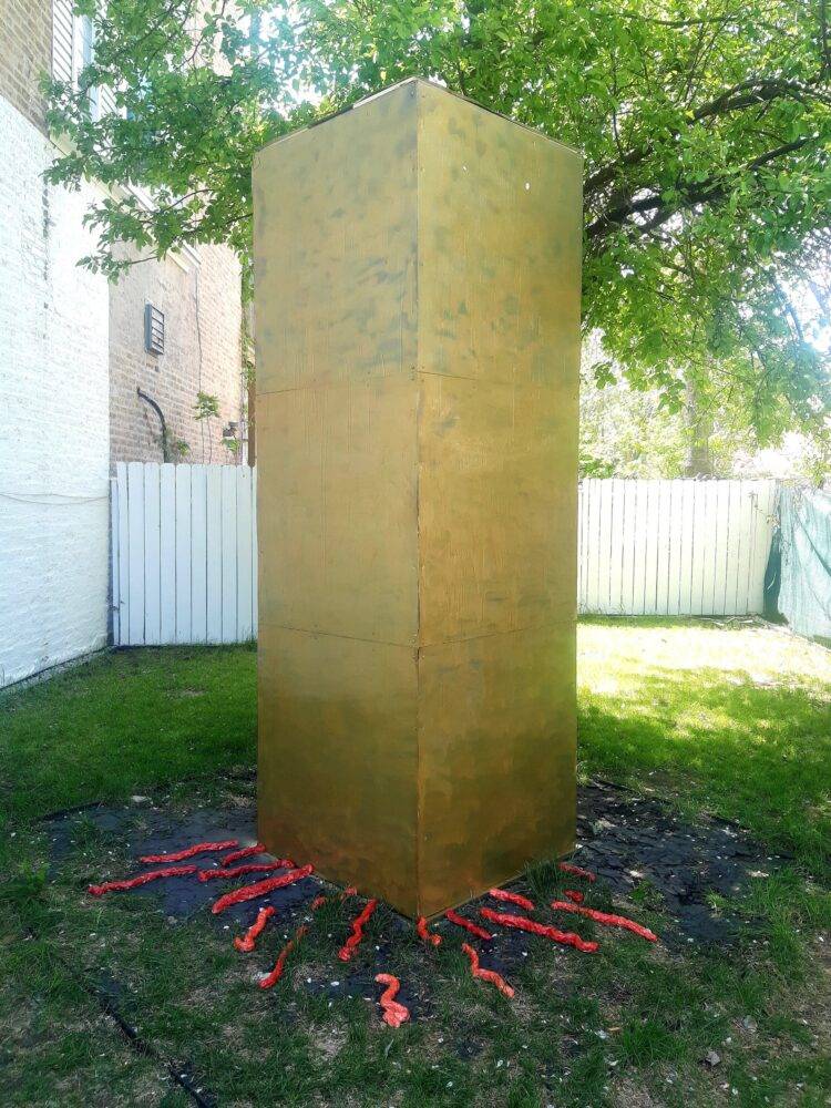 A brown-gold monolith stands in a grassy backyard. Red veins leak out from the bottom of the monolith.