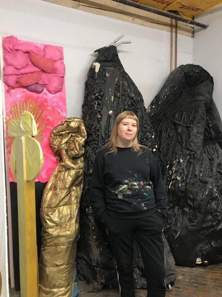 A woman, the artist Sonya Bogdanova, wears all black and strands in front of black, gold and pink works inside her studio.
