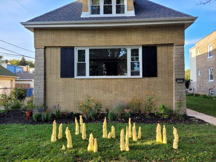 Yellow stalagmites made by Bogdanova sit on a green lawn in front of a beige house.