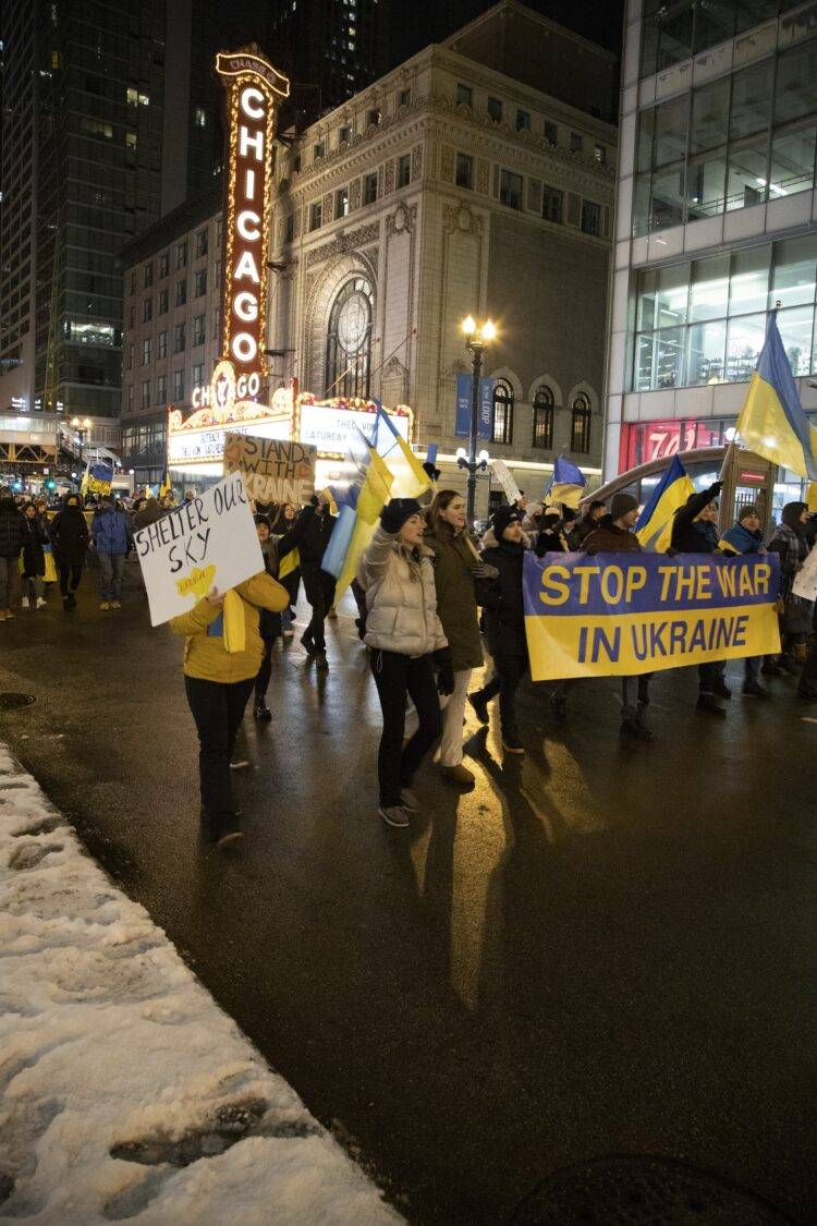 A crowd of people marches in front of the Chicago Theater sign. The people are holding Ukrainian flags and are holding a sign that reads: "STOP THE WAR IN UKRAINE."