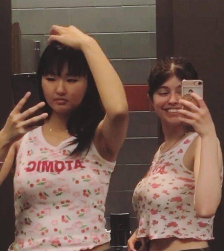 Two girls take photos in a mirror, both wearing white-and-pink tank tops with the word "ATOMIC" in all-capital letters.