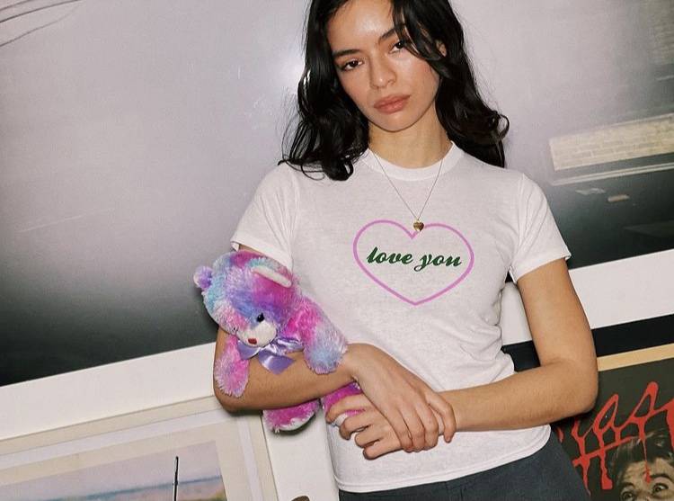 A model holding a pink-blue-purple bear wears a white tee shirt featuring the words "love you" in cursive, ensconced in a pink heart. 