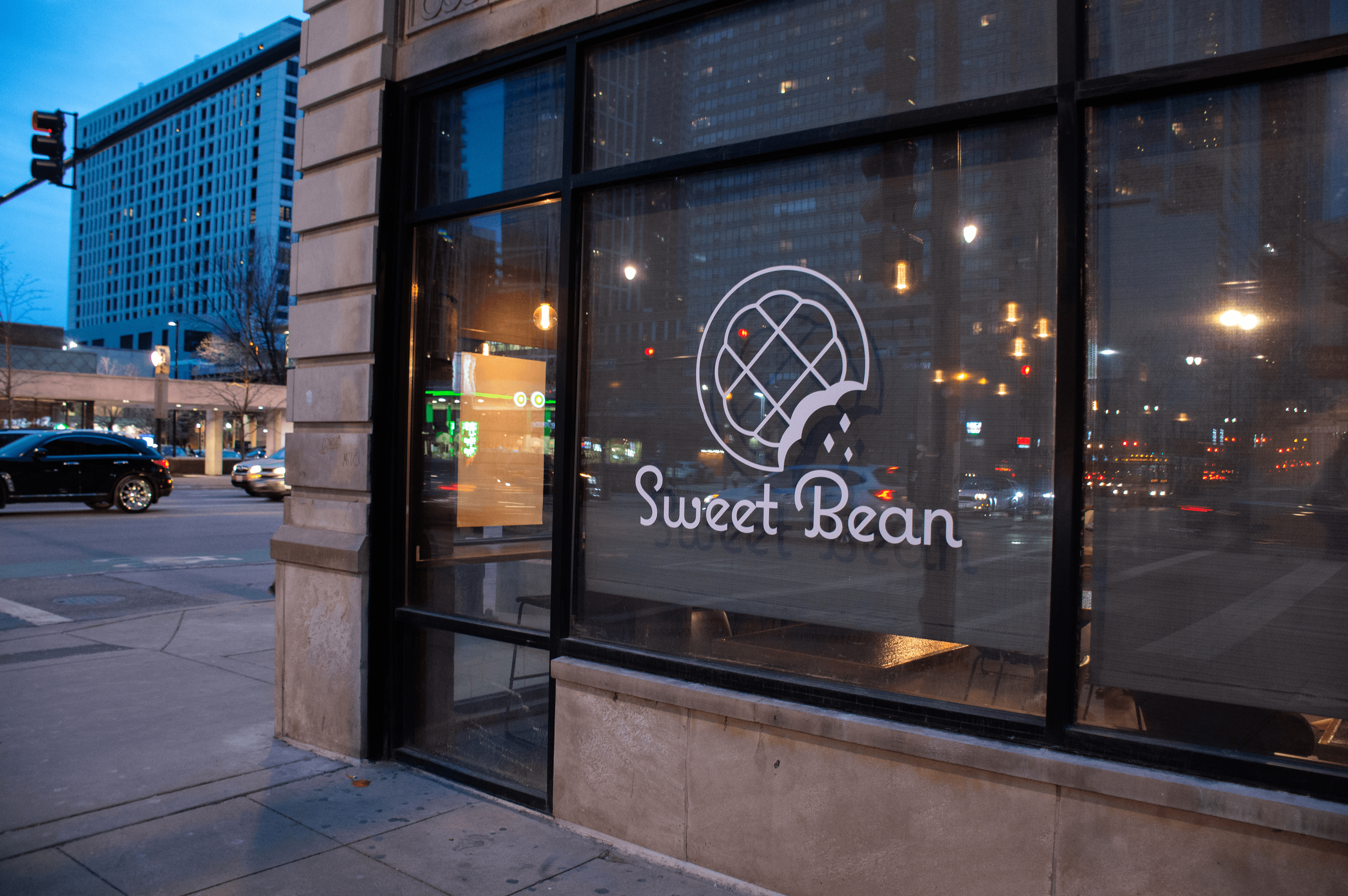 Store front with reflection of the city in the glass and “Sweet Bean” written in big letters under a graphic of a crumbled cookie.