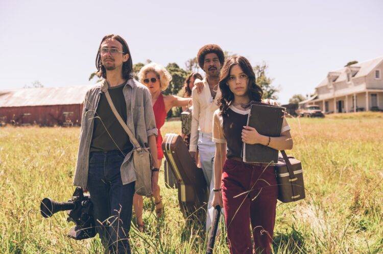 Five young people walk through a field. A brown haired woman and a long, dark haired man are leading the group. The woman clutches a notebook against her chest, the man is carrying a professional looking video camera.