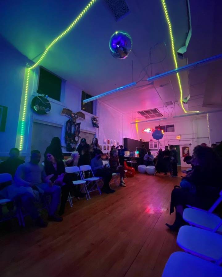 A narrow room with wooden floors. Green and blue neon lights are strung aross the walls, in the back is a glowing pink light. A disco ball hangs from the ceiling.