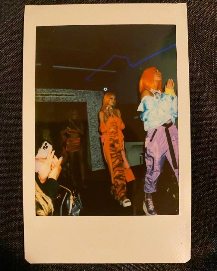 A photograph of a polaroid image. In the polaroid are two models walking with orange wigs on. One has their arms up, as through mid-clap. The other wears a floor length dress and is glancing away from the camera.