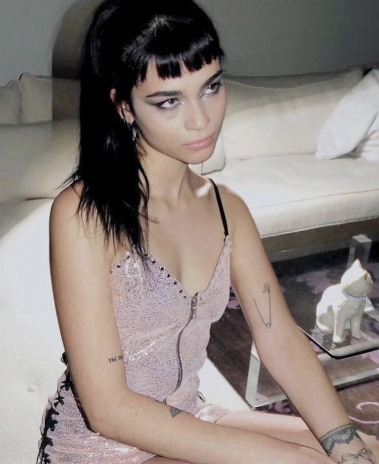 A woman with dark hair and faint tattoos poses in a pale pink dress.