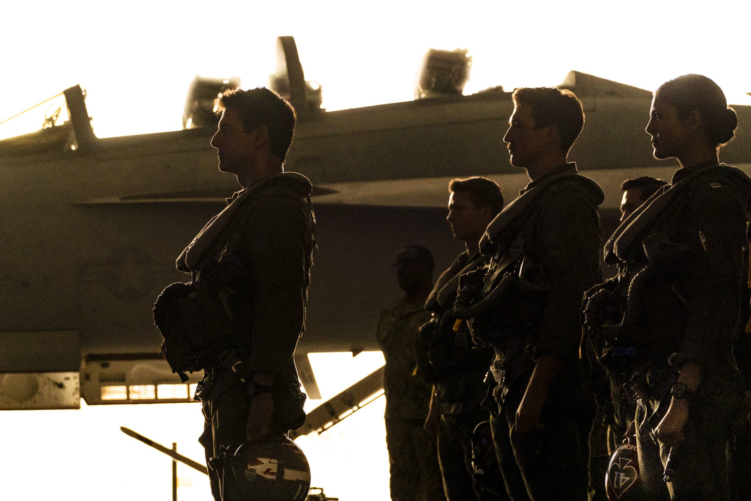 Silhouettes of five fighter pilots from the side. Tom Cruise stands in front, the other four behind him. There is a jet in the background.