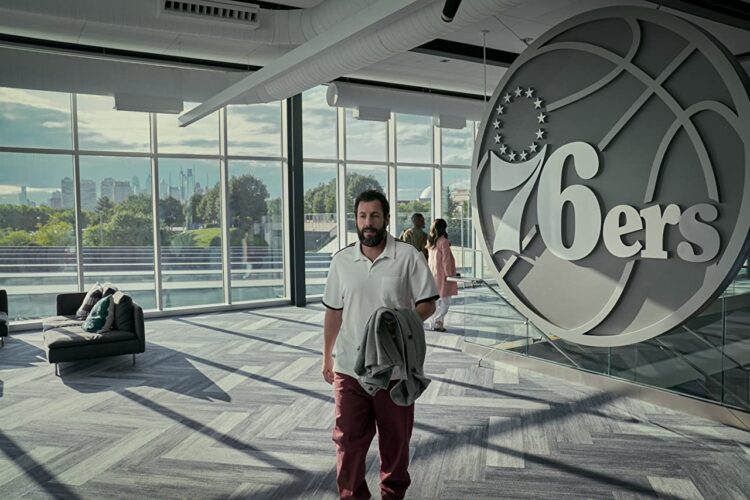 Film still from the movie Hustle. Adam Sandler walking toward the camera with a contemplative look on his face. In the background is a large basketball logo for the Philedelphia 76ers, and a wall of windows. 