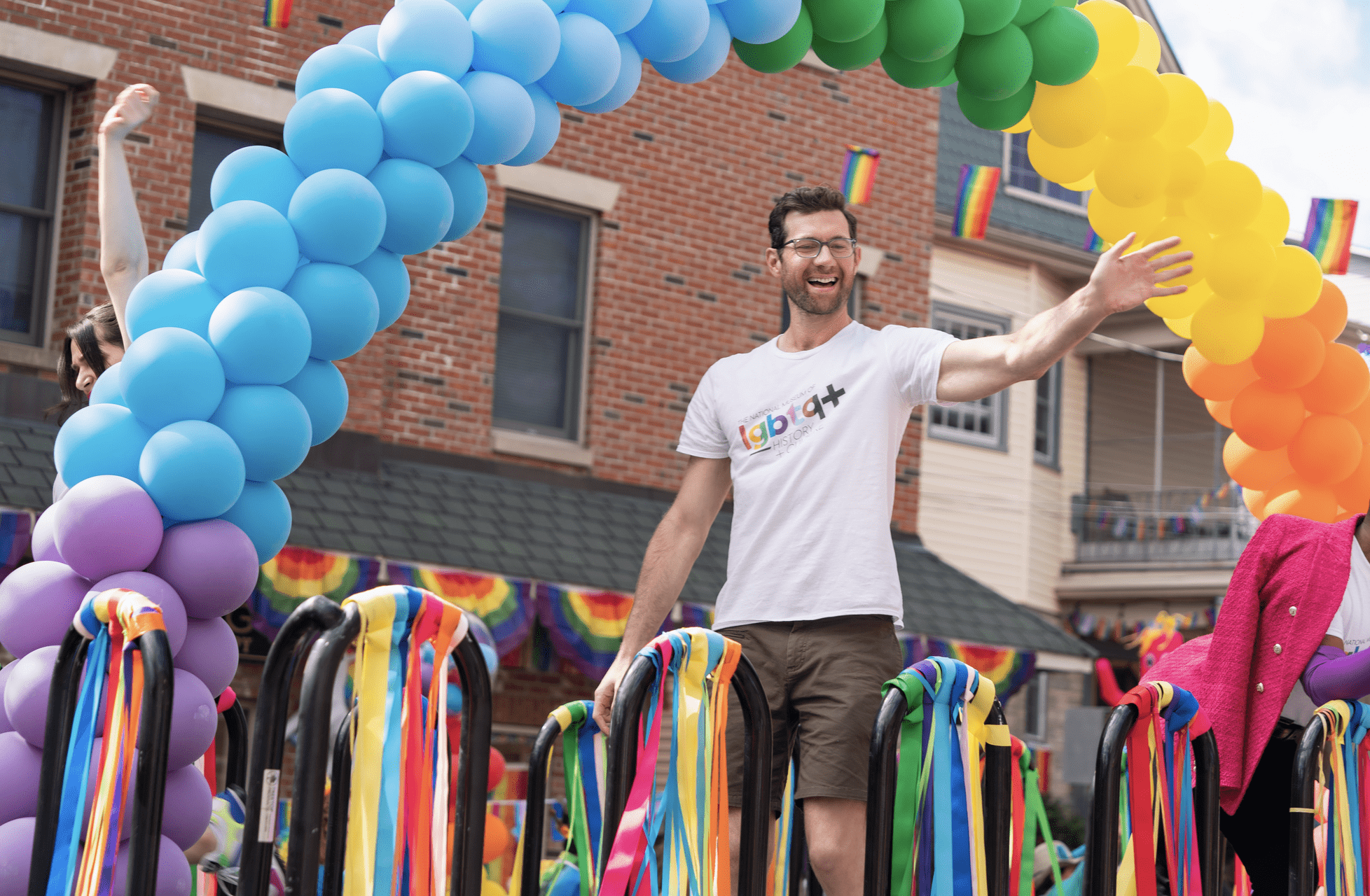A photo of Billy Eichner in the movie "Bros" standing and waving on a Pride float.