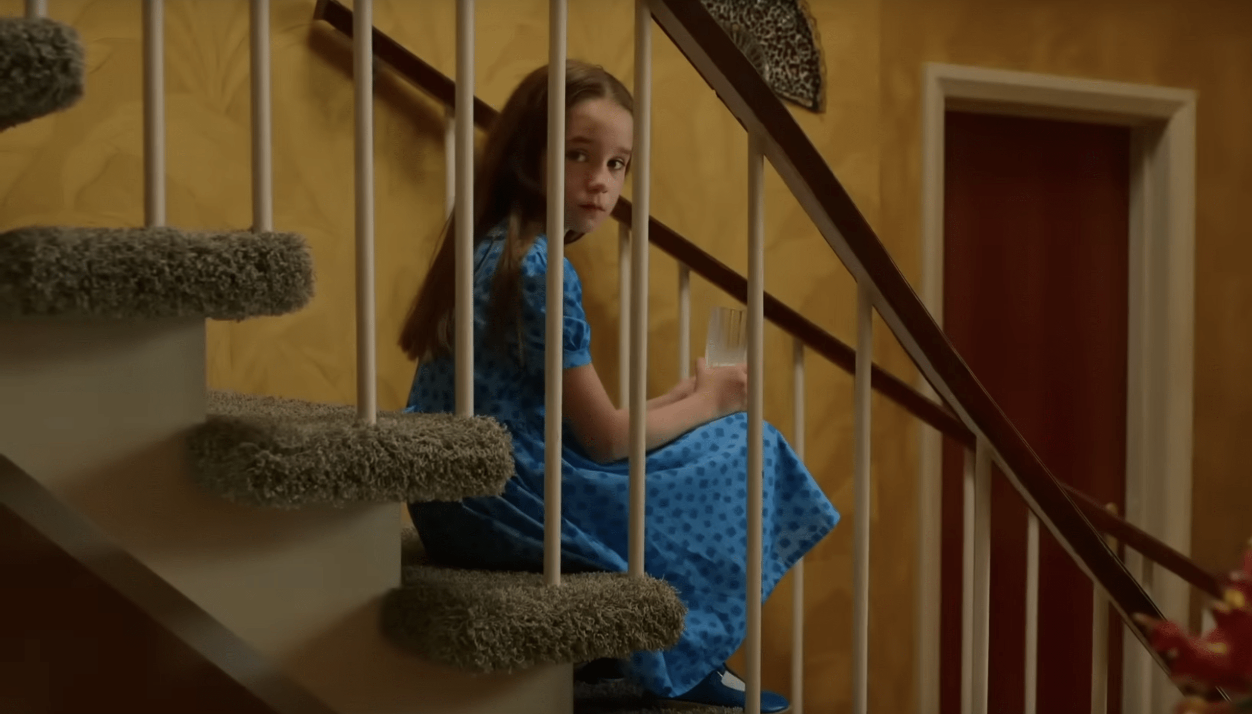 A small girl in a blue dress sitts on a set of carpeted stairs. She's looking over her shoulder sadly.