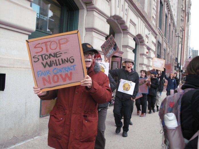 IMG_0871 A protester at AICWU’s informational picket holds a sign that says “STOP STONE-WALLING FAIR CONTRACT NOW.”